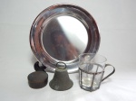 Quatro peças diversas em metal espessurado a prata: a) Caneca com estrutura em metal, interior com copo de vidro, manufatura Nassau. Alt. 9cm. b) Caixinha redonda procedente da Cordilheira dos Andes, Chile. 2 x 6cm. c) Sineta em metal amarelo. Alt. 7cm. d) Bandeja em inox, diâm. 24cm.