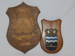 Duas placas em madeira emoldurando escudos de instituições marítimas, sendo uma com escudo em madeira entalhada do Comando Geral da Armada Uruguaia, 38 x 25cm; e um com escudo não identificado em resina, 25 x 16cm.
