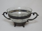 Centro de mesa, base em metal prateado, 4 pés em garras e ornados com caras de leão, alças em volutas, bowl interior em vidro lapidado com frisos. Marcado Majedi, São Paulo. 13 x 28cm.