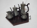 Conjunto para serviço de chá e café em estanho nacional, composto por 5 peças: bule para chá, bule para café, leiteira, açucareiro e bandeja de madeira. Decoração por frisos, alças e pegas em madeira, bandeja sextavada em madeira, alças em estanho da mesma manufatura. Pega da tampa do açucareiro quebrada. Alt. maior 24cm, bandeja diâm. 42cm.