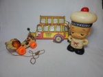 Três peças infantis: a) Carrinho de puxar na forma de cão em madeira policromada. Comp. 21cm. b) Boneco decorativo em plástico representando marinheiro. Alt. 31cm. c) Placa decorativa na forma de ônibus "Koo Koo Munga", 40 x 26cm.