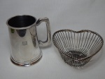 Duas peças em metal prateado: a) Caneca lisa com símbolo cinzelado. Manufatura Eberle. Alt. 13cm. b) Cesta em aramado prateado na forma de coração, 7 x 15 x 15cm.