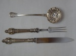 Três peças de servir em metal espessurado a prata: a) Trinchant para assados, cabo em alpaca ricamente ornamentada por acantos e volutas. Comps. 27 e 31cm. b) Concha polvilhadora com vazados, decorada com acantos. Manufatura Hart. Comp. 21cm.