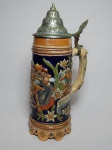 Tankard com caixa de música em cerâmica vitrificada alemã, decoração moldada na pasta policromada com cenas típicas alemãs, tampa em metal prateado. Caixa de música ao fundo, toca a música folclórica alemã "O Mein Papa", porém sem funcionamento aparente. Alt. 30cm.