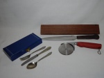 Cinco peças de serviço diversas: a) Colher, garfo e faca no estojo, cabo em alpaca, lâmica em inox. Manufatura Wolff. Comp. maior 17cm. b) Faca "cuts-all knife", manufatura Burnco, cabo em madeira, aplicado com a marca do "The Tilden Bank", Nebraska, Estados Unidos. Comp. 33cm. c) Cortador de pizza em inox, manufatura Wilton. Comp. 21cm.