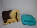 Duas peças de cozinha: a) Sanduicheira elétrica Kitchen Selectives, modelo SW-22TQ, 120V. Não testada e sem garantia. 8 x 24 x 24cm. b) Cortador de frios manual, estrutura em plástico. 20 x 28 x 20cm.