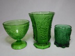 Três peças em vidro verde da década de 50: a) Vaso moldado com cachos de uvas e folhas de parreira, base e corpo oitavados. Alt. 16cm. b) Taça moldada com flores, frisos com gregas. Alt. 12,5cm. c) Copo moldado com acantos e volutas. Alt. 9,5cm.