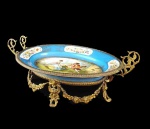 SEVRES  CENTRO DE MESA EM PORCELANA COM GUARNIÇÃO EM BRONZE. FEITIO OBLONGO, FUNDO NA TONALIDADE AZUL CELESTE. ABA DECORADA COM RESERVAS FLORAIS EM ESMALTE REMATADAS POR GUIRLANDAS EM OURO. CALDEIRA COM CENA GALANTE. GUARNIÇÃO EM BRONZE ORMOLU. PEGAS COM FEITIO DE ALÇAS DUPLAS.  FRANÇA, SEC. XIX. 24,5 CM DE COMPRIMENTO.