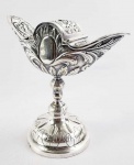LINDA NAVETA EM PRATA DE LEI. . ELEGANTE FEITIO COM DECORAÇÃO RELEVADA DE ELEMENTOS FLORAIS, FOLHAS DE ACANTO E ROCAILLE. BRASIL, SEC. XIX. 18 CM DE ALTURA. NOTA:  A naveta é objeto que porta o incenso. Normalmente em forma de barca, de onde vem seu nome. Através de uma colherinha, se deita o incenso no turíbulo. O uso do incenso é antiquíssimo, anterior à própria vinda de Cristo. As primeiras menções ao seu uso provavelmente vem dos egípcios, no III milênio a.C.. Seu uso sempre foi relacionado à aromatização e purificação do ambiente e a uma oferta de odor agradável à divindade. Os hebreus, por exemplo, utilizavam amplamente o incenso no Templo de Jerusalém, onde havia o altar do Incenso, no qual era queimado continuamente o incenso em tributo a Deus. (Cf. Ex 30; Lv 16,12). Talvez por ser amplamente utilizado entre os pagãos, a Igreja nos seus primeiros anos evitava o seu uso dentro da Liturgia, para fazer uma distinção clara entre a liturgia pagã e a liturgia cristã, em particular porque o incenso oferecido aos deuses pagãos era uma grande traição à fé, e vários santos foram torturados e martirizados por se recusarem a fazê-lo. (Como S. Cassiano, S. Policarpo, S. Dinis, S. Cristina, S. Jorge e muitos outros). Uma vez que foi destruído o paganismo, a partir do século IV o incenso começou a ganhar um espaço maior na Liturgia, sobretudo nas dedicações do altar. A primeira igreja a receber o incenso foi a igreja do Santo Sepulcro, e então o uso foi se espalhando. A primeira aparição formal do incenso em Roma é do século VII, como gesto de honra ao papa e ao Evangeliário. Dentro da Missa, o incenso começou a ser utilizado por volta do século IX, no início da Missa, de modo similar como é hoje: Altar, clero, oblatas. Isto se fazia se passando o incensário (Uma espécie de vaso onde se queimava o incenso) à frente dos objetos e das pessoas. A liturgia foi se desenvolvendo até chegar ao modo como temos hoje. Foi assim que na idade média a naveta começou a ganhar uma forma de navio. Isto ao mesmo tempo significada duas coisas: A primeira é a lembrança da Igreja como a Barca de Pedro. A segunda é que a vida espiritual deve ser sempre um caminho, daqui às Mansões Eternas. O navio simboliza esta mobilidade.