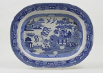 STAFFORDSHIRE  LOUÇA DE SALGUEIRO OU POMBINHO - GRANDE TRAVESSA EM FAIANÇA DECORADA EM FAIANÇA NA TONALIDADE AZUL COM REPRESENTAÇÃO ALUSIVA A LENDA DO SALGUEIRO DE ORIGEM ORIENTAL. INGLATERRA, SEC. XIX. 34 CM DE COMPRIMENTONOTA: A lenda do salgueiro ou pombinho ganhou notoriedade a partir de meados do sec. XIX quando passou a ser usada com frequência em louças inglesas produzidas pela manufatura Stafordshire desde 1868. A lenda oriental resumida é a seguinte: Havia uma bela jovem filha de um abastado mandarim que morava em seu palácio. A jovem enamorou-se de um rapaz de origem humilde contrariando os projetos do pai que desejava um casamento que trouxesse vantagens a sua casa. Para coibir a relação dos apaixonados, a moça foi mantida por anos em uma pequena casa no fundo do palácio do mandarim. Ali ela percebia a passagem do tempo pelo florescimento de um grande salgueiro que domina a cena representada na louça. Certo dia a jovem recebeu em uma casca de coco envia