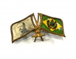 DOM LUIZ DE ORLEANS E BRAGANÇA  O PRÍNCIPE PERFEITO   RARO  PIN DE PROPAGANDA MONARQUISTA DA SECADA DE 1910 COM FEITIO DE DUAS BANDEIRAS HASTEADAS SENDO UMA A DO IMPÉRIO DO BRASIL E OUTRA COM RETRATO DE  DOM LUIZ DE ORLEANS E BRAGANÇA, CHEFE DA CASA IMPERIAL E HERDEIRO PRESUNTIVO DO TRONO BRASILEIRO. EUROPA, DEC. 1910. 4 CM DE COMPRIMENTO.NOTA: Luís de Orléans e Bragança (Petrópolis, 26 de janeiro de 1878  Cannes, 26 de março de 1920), foi o segundo filho da princesa Isabel do Brasil e do príncipe consorte Gastão de Orléans, Conde d'Eu, e neto do último imperador do Brasil, Pedro II. Príncipe do Brasil até novembro de 1889, tornou-se pretendente ao extinto trono imperial brasileiro em 30 de outubro de 1908, quando o seu irmão, Pedro de Alcântara, renunciou a seus direitos dinásticos. É o patriarca do chamado ramo de Vassouras da família imperial brasileira. Batizado Luís Maria Filipe Pedro de Alcântara Gastão Miguel Rafael Gonzaga de Orléans e Bragança, desde pequeno revelou possuir uma personalidade forte e determinada, como quando, em viagem à Europa com sua família no dia 23 de fevereiro de 1887, em que ocorreu um terremoto logo no amanhecer e enquanto seu irmão mais velho Pedro ficara nervoso e chorava, Luís simplesmente ficou impassível, como se a situação pouco o afetasse. As discrepâncias entre ele e o irmão mais velho eram notórias, como seu pai descreveu em uma carta datada de fevereiro de 1889 onde revelou Pedro como "tão incapaz e descuidado nisso (jogar bilhar com dom Pedro II) quanto em tudo o mais". Enquanto Pedro era gentil e simpático, não gostava de estudar e se revelava normalmente desajeitado, Luís tinha força de vontade, era muito altivo e perspicaz. Gaston, o conde d´Eu, afirmou em uma carta em 1890 que o "Bebê Pedro sempre se destaca pela indolência e a inépcia, ao passo que "Luís faz exatamente o mesmo trabalho escolar sozinho, com um prestígio e uma capacidade admiráveis". O príncipe muito cedo revelou interesse pelas letras que, ao se tornar adulto, faria-o dedicar-se a escrever diversas obras que mais tarde publicou relatando suas experiências de viagens: Dans les Alpes, Tour d´Afrique, Onde quatro impérios se encontram, Sob o Cruzeiro do Sul.Luís, de "natureza irrequieta, a necessidade e ação que, nos anos juvenis, o impelia a esportes impulsionou-o, na maturidade, à ação política".  Não sendo a toa que no auge da campanha abolicionista, ele e seus irmãos publicavam um jornal abolicionista no palácio de Petrópolis. Com a Proclamação da República, em 15 de novembro de 1889, a princesa Isabel preferiu enviar os filhos para Petrópolis, onde mais tarde Luís recordou que "encerrados no palácio, deixaram-nos durante dois longos dias na mais completa ignorância do que se passava lá fora", até que foram entregues de volta aos seus pais e partiram para o exílio forçado Como não puderam levar nada, a não ser alguns objetos pessoais de mão, a família imperial se viu numa situação financeira muito complicada, que piorou com a recusa de dom Pedro II de cinco mil contos de ajuda de custo oferecidos pelos golpistas.Tiveram que se contentar com a ajuda de amigos e até mesmo do pai de Gaston, conde d´Eu.  Resolveram fixar-se nos arredores de Versalhes em 1890, quando Pedro tinha quinze anos, Luís treze e o irmão mais novo, Antônio, apelidado de "Totó", nove. Apesar dos mais variados esforços dos monarquistas no Brasil para ressuscitarem a monarquia, após a morte de dom Pedro II em 1891, nenhum membro da família imperial colaborou com nenhum tipo de ajuda, nem mesmo com palavras de apoio explícito. Pedro, irmão mais velho de Luís e herdeiro da princesa Isabel do Brasil e, portanto, do extinto trono imperial do Brasil, tornou-se maior de idade em 1893, mas não possuía capacidade e muito menos desejo para assumir a causa. No mesmo ano Pedro partiu para Viena, capital do então Império Austro-Húngaro para estudar na escola Militar de Wiener Neustadt, pois segundo sua própria mãe, "é preciso que faça alguma coisa e a carreira militar nos parece a única que ele deve seguir". Luís e seu irmão mais novo, Antonio, logo o seguiram para a mesma escola militar. Em 1896 Pedro conheceu uma moça chamada Elizabeth de Dobrzenicz e logo se apaixonaram, tendo o casal combinado bastante em termos de temperamento e caráter. Enquanto isso, Luís Maria Filipe Pedro de Alcântara Gastão Miguel Rafael Gonzaga de Orléans e Bragança, "era um ativista; ambicioso e voluntarioso, encarava o mundo como algo a ser conquistado. Praticante de alpinismo, escalou o Mont Blanc em 1896. A uma visita ao sul da África, seguiu-se uma longa e ousada excursão à Ásia Central e à Índia. Sobre essas três experiências ele escreveu e publicou". Não sendo a toa que era justamente Luís, que dona Isabel e o conde d´Eu viam a única pessoa entre os membros de sua família capaz de manter a causa monárquica no Brasil. Ao retornar de suas aventuras em 1907, Luís planejou um projeto ambicioso que seria desafiar o decreto de banimento da família imperial, viajando para o Rio de Janeiro. Sua súbita chegada criou um rebuliço na antiga capital imperial, tendo sido amplamente "noticiado nos jornais, o episódio alcançou grande repercussão nos meios políticos, colocando a família imperial no centro das atenções e muitos monarquistas e curiosos vieram recebê-lo".No entanto, Luís foi impedido de desembarcar e não foi permitido pisar em sua terra natal pelo governo republicano. Inclusive enviou um telegrama a sua mãe dizendo: "Impedido de desembarcar pelo governo, saúdo da baía da Guanabara, na véspera do 13 de Maio, a redentora dos cativos." Algum tempo depois, relatou as experiências dessa viagem em Sob o Cruzeiro do Sul, publicado em 1913. Em 1908, Luís ficou noivo de uma prima, Maria Pia de Bourbon, sobrinha-neta de sua avó materna, Teresa Cristina de Bourbon-Duas Sicílias, enquanto o seu irmão mais velho, Pedro, herdeiro da princesa Isabel, desejava casar-se com Elizabeth de Dobrzenicz. Tal casamento não seria permitido pela então chefe da casa imperial, a princesa Isabel, pois Elizabeth, ou "Elsi", como era chamada, embora nobre não fazia parte de nenhuma família reinante da Europa, mesmo que alguma deposta. A princesa Isabel, como mãe, não desejava o sofrimento do filho, e concordou com o casamento de Pedro contanto que ele renunciasse ao seu direito ao trono. Pedro, que não possuía interesse em tornar-se imperador, assinou a renúncia no dia 30 de outubro de 1908. Pedro renunciou solenemente, assinando um documento reconhecido por sua mãe. O casamento de Luís com Maria Pia foi celebrado em 4 de novembro, e o do Pedro com Elizabete, dez dias depois.1 Do matrimônio de Luís com Maria Pia nasceram três filhos: dom Pedro Henrique (1909-1981), que se tornou o sucessor direto da princesa Isabel e Chefe da Casa Imperial após o falecimento desta em 1921; Luís Gastão (1911-1931) e Pia Maria (1913-2000). A princesa Isabel não tardou a manifestar sua opinião quanto aos netos, escrevendo em 1914 uma carta dizendo: "envio-lhe uma fotografia minha com meus netos do Luís. Pedro Henrique cada vez se desenvolve mais e é criança inteligentíssima. Os avós têm um amor especial pelos queridos netinhos". Com a renúncia do irmão, Luís pôde finalmente colaborar efetivamente com o movimento monarquista brasileiro, assumindo claramente sua posição como herdeiro do trono (após sua mãe) e buscando assumir a liderança da campanha restauradora. Seu esforço para reverter todo o mal causado pela inércia da família imperial quanto à causa monárquica foi de grande valia, e em 1909 apresentou um manifesto político aos monarquistas brasileiros que tinha por objetivo retomar a campanha que estava estagnada há alguns anos. Seu intentou logrou sucesso, pois conseguiu reunir correligionários em diversos estados do Brasil. Algumas das cartas do príncipe revelam seus planos de restauração, como a escrita para Martim Francisco de Andrada III. "quanto me custa ficar aqui, de braços cruzados, quando penso que um punhado de homens decididos bastaria para arrancar a Pátria das garras dos aventureiros que a exploram." "Ainda não sei qual será a sua atitude na questão das candidaturas. Quanto a mim, julgo ambos os candidatos 'indesejados'; mas a ter de optar, optaria pelo Rui Barbosa, cujos partidários representam o elemento mais são e de maior prestígio no país. Parece-me mesmo que poderíamos aproveitar o momento para um acordo com os próceres desse grupo, a fim de conseguirmos um esforço comum pela restauração, logo após as eleições presidenciais. Que lhe parece?" O príncipe atuou de 1907 até 1920, e defendia o federalismo, o serviço militar obrigatório e uma melhoria na qualidade de vida dos operários. No primeiro caso, era a favor de uma maior descentralização e liberdade política e econômica para os estados brasileiros. No segundo, refutava o antigo costume de alistamento das forças armadas baseado em indivíduos provenientes das camadas sociais marginalizadas em favor de uma força militar verdadeiramente profissional formada por elementos de toda a sociedade. O terceiro e mais importante caso era a pregação de uma monarquia amparada numa legislação social que possibilitasse melhores condições de vida para os operários brasileiros. Em carta expôs seus pensamentos: "Quanto a nós, monarquistas, devemos convencer o operário da verdade de que, no caso de uma restauração, a sua situação só poderia melhorar." Luís defendia ideias muito a frente de seu tempo e a necessidade de garantir condições dignas de subsistência para os trabalhadores brasileiros seria observada somente trinta anos depois na ditadura de Getúlio Vargas. No início do século XX, tanto o governo quanto os políticos brasileiros sequer admitiam a possibilidade de existência de direitos básicos como férias, greve, horas máximas semanais de trabalho, entre outros. A visão progressista de Luís o fazia ser acusado de "radical" e "socialista" quando na realidade seu intento era justamente impedir a adesão do operariado ao socialismo, comunismo ou mesmo ao anarquismo. Em agosto de 1914 se iniciou a Primeira Guerra Mundial, que na época foi conhecida como "A Grande Guerra". A invasão da França pela Alemanha "ofereceu uma válvula de escape tanto para o idealismo como para o ativismo de Luís, que segundo suas próprias palavras, era um 'soldado no fundo do coração'. Ele e o irmão Antônio precipitaram-se a defender a pátria dos ancestrais". Como a lei os proibia de servir nas forças da nação, por serem membros da família real francesa, ambos se alistaram como oficiais do exército inglês. Em 1915, combatendo nas trincheiras de Flandres e servindo como oficial de ligação, Luís contraiu um tipo agressivo de reumatismo ósseo que o deixou debilitado e incapaz de andar Foi retirado em estado grave das trincheiras e levado para segurança, a fim de poder recuperar-se da moléstia. Em consequência de suas ações no conflito e por sua bravura, Luís recebeu diversas condecorações: Medalha Militar do Yser, pelo rei Alberto I da Bélgica; Legião de Honra, no grau de cavaleiro, e a Cruz de Guerra, pelo governo francês; a British War Medal, a Victory Medal e Star pelo Reino Unido da Grã-Bretanha.1 A grave doença contraída nas trincheiras resistiu a todas as formas de tratamento e sua saúde foi piorando cada vez mais, até que a morte o levasse em 26 março de 1920. Em 1918 foi publicado o Arquivo Nobiliárquico Brasileiro, obra pioneira que trata da nobiliarquia brasileira. Seus autores, os luso-brasileiros Rodolfo Smith de Vasconcelos, segundo barão de Vasconcelos, e seu filho, Jaime Smith de Vasconcelos, terceiro barão de Vasconcelos, dedicaram a obra a sua alteza imperial o Senhor Dom Luiz de Orléans Bragança. Príncipe desconhecido pelos brasileiros atualmente, tendo sido um exemplo de cavalheirismo e seu amor por sua terra natal foi demonstrado em todos os momentos possíveis, ainda mais depois que assumiu a posição de herdeiro da mãe em 1908, envolvendo-se publicamente na campanha de restauração do trono no Brasil e tomando parte ativa nos movimentos monarquistas até a Primeira Guerra Mundial. Suas ideias inovadoras, como a inclusão da questão social com maiores direitos à classe operária e melhor qualidade de vida para os brasileiros na agenda política, numa época em que era considerada "caso de polícia" pelos governantes da República Velha, lhe renderam o epíteto de "príncipe perfeito" ou mais precisamente como rei Albert II dos Belgas falou a seu respeito: "homem como poucos, Príncipe como nenhum".