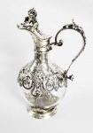 LINDO DECANTER EM CRISTAL COM GUARNIÇÃO EM PRATA DE LEI. MARCAS  DE CONTRASTE PARA FRANÇA, FINAL DO SEC. XIX. TAMPA TEM PEGA COM FEITIO DE DRAGÃO HERÁLDICO SEGURANDO PERGAMINHO. TERÇO SUPERIOR E BASE REVESTIDOS EM PRATA DE LEI COM DELICADAS ROCAILLES E VOLUTAS FORMANDO GROTESCO COM CABEÇA DE ANIMAL. ALÃ TAMBEM TEM FIGURAS GROTESCAS. SENDO UM HOMEM VERDE E UMA FERA. FRANÇA, FINAL DO SEC. XIX. 23 CM DE ALTURANOTA: O termo grotesco deriva da palavra italiana grottesco (de gruta, ou cova), surgindo como um estilo artístico inspirado nas decorações da Roma Antiga, descobertas em ruínas escavadas durante o Renascimento. Esses monumentos subterrâneos eram conhecidos por grottes, e foram encontrados em 1480, num local que hoje é conhecido como Oppius. Essas escavações revelaram os restos do palácio Domus Aurea, erguido por Nero após o grande incêndio que destruíra parte de Roma no ano de 64. O palácio possuía uma espécie de pintura ornamental totalmente incomum, principalmente em relação à imagem que se tinha do estilo classicista de arte romana. Esse estilo recém descoberto era baseado em formas, como figuras delirantes, máscaras e animais. O ornamento grotesco, geralmente, se caracteriza pela criação de universos fantasiosos, cheios de seres humanos e não-humanos, fundidos e bizarros. As visitas às grutas se tornaram uma verdadeira viagem insólita, em que uma nova noção de arte se fazia a partir do estranho, do irreal e do assustador. Diversos pintores fizeram excursões regulares às ruínas da Domuns Aurea, entre eles Giovanni da Udine, Pinturicchio e Rafael Sanzio, principalmente para conhecerem os ornamentos criados pelo pintor Fâmulo (também conhecido como Fabullus, ou Fábulo). Um dos primeiros exemplares do ornamento grotesco é o friso da Anunciação, feito por Carlo Crivelli, em 1486, que chama atenção pela deformação de elementos naturais. As pinturas grotescas feitas por Luca Signorelli, entre 1499 e 1504, para a Catedral de Orvieto, mostram uma confusão de elementos e animais estranhos, usados para representar a Divina Comédia, de Allighieri. Entre os mais conhecido e influentes obras grotescas, encontram-se as obras de Rafael Sanzio, para o forro e pilares das loggie do Vaticano, de 1515, formados por arabescos e linhas ondulados e verticais, com animais e vegetais entrelaçados. O grotesco como um estilo marginal, até que a grande difusão do estilo que se deu já no fim do século XVI, quando já tomava grande parte arquitetura italiana. Muitos outros artistas renomados passaram a realizar suas obras com base no estilo grotesco, como Gaudenzio Ferrari, Signorelli, Filippino Lippi, Andrea di Cosimo, Giuliano da Sangallo e até mesmo Michelangello. Logo após essa explosão no país, o estilo começou a entrar em outros países da Europa. Rapidamente a noção de grotesco perde o sentido técnico, especificamente, como um tipo de arte romana e um estilo renascentista, transformando-se em um adjetivo para indicar aquilo que é bizarro, fantástico e extravagante. Posteriormente, o movimento neoclássico, ao resignar a linha curva e retorcida dos estilos barroco, acaba por rejeitar, de modo geral, o estilo grotesco, que é considerado excessivamente despropositado, colocando-o novamente na marginalidade. Acaba sendo muito valorizado pelos românticos, para os quais a arte deveria representar tanto o que é belo e admirável, quanto o que é feio e deformado. Esses artistas transformam o grotesco numa espécie de categoria estética e literária, para fazer referência a um tipo de descrição ou de tratamento deformador da realidade, que pode ter como objetivo provocar gargalhadas, com uma intencionalidade satírica. Se tomarmos o grotesco em uma concepção mais ampla, com seus recursos de deformação de figuras humanas, com uma definição dramática, podemos encontrar o grotesco na arte brasileira desde as obras de Aleijadinho. Atualmente, a palavra grotesco apanhou o sentido de adjetivo de sentido bizarro ou ridículo, sem muita relação com as pinturas que originaram o termo.