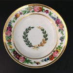 DOM PEDRO I - SERVIÇO DA ILÚSTRISSIMA CÂMARA OU SERVIÇO FUMO E CAFÉ  RARO PRATO EM PORCELANA COM ABA DECORADA COM LUXURIANTE GUIRLANDA COM FLORES COLORIDAS. CALDEIRA DE RAMO DE FUMO E CAFÉ UNIDOS POR CAPRICHADO LAÇO. FOI ENCOMENDADO PELA SENADO DA CORTE PARA SER OFERTADO AO IMPERADOR DOM PEDRO I, QUE EM 1823 CONFERIU AO PARLAMENTO CARIOCA A HONRARIA DO TÍTULO DE ILUSTRÍSSIMA CÂMARA MUNICIPAL(VIDE NOS CRÉDITOS EXTRAS DESSE LOTE O ANTIGO PRÉDIO DA ILUSTRISSIMA CÂMARA DO RIO DE JANEIRO NA ÉPOCA DE DOM PEDRO I). O ANÚÁRIO DO MUSEU IMPERIAL BRASILEIRO PUBLICADO EM  1942 TRAZ SOBRE ESSE SERVIÇO, AS SEGUINTES INFORMAÇÕES:  Ainda desse tempo um belíssimo serviço, também raro nas nossas coleções. É o que pertenceu a ilustríssima Câmara Municipal do Primeiro Reinado, todo branco, borda em cercadura de flores, ramos de fumo e café ao centro. Pinturas a encarnado e verde, lembrando muito a decoração da fábrica Dagoty, têm eles, entretanto, a marca de De Roche 12. Ao que já ouvi dizer de pessoa informada, esse aparelho foi, quase em vésperas da abdicação, presenteado pelo imperador a uma afilhada de batismo, de família afeiçoada ao paço e moradora no caminho velho de Botafogo 13. Em seguida, dispersou-se pelos leilões e antiquários, alcançando os museus e as mãos ciosas dos felizardos que possuem restos dele. MARCA DO DECORADOR DEROCHE NO VERSO. PEÇA IDÊNTICA INTEGRA O ACERVO DO MUSEU IMPERIAL EM PETRÓPOLIS E ENCONTRA-SE REPRODUZIDO A PÁG. 207 DO LIVRO "LOUÇA DA ARISTOCRACIA NO BRASIL", POR JENNY DREYFUS. INÍCIO DO SÉCULO XIX. 22 CM DE DIAMETRO. NOTA: A Câmara Municipal foi desde sempre um órgão prestigiado na administração portuguesa e também na de suas colônias. Era destinada aos cidadãos proeminentes da sociedade. No Rio de Janeiro, sede do Reino Unido nos tempos de Dom João VI e sede da Corte do Império a partir de Dom Pedro I, tão mais importante foi a Câmara do Senado Municipal. Tal foi que em 1823 nas festividade de aniversário do célebre Dia do Fico, o primeiro passo para nossa Independência no ano seguinte, o Imperador  achou por bem galardoar a Câmara com o título de ILUSTRÍSSIMA CÂMARA MUNICIPAL DO RIO DE JANEIRO A distinção dos vereadores já se dava com as vestimentas. Havia um traje especial desde as ordenações da restauração de Portugal. No auto de levantamento e juramento do d. João IV, a 1o de dezembro de 1640, lê-se que o presidente da Câmara de Lisboa, conde de Castanhede, compareceu vestido de veludo negro aforrado de tela branca e mangas da mesma cor. No Rio de Janeiro, o traje dos vereadores durante a permanência da corte portuguesa no Brasil e sob o governo de d. Pedro I, até 1828, era composto de casaca, calção e colete pretos, meias brancas e sapatos de fivela. O chapéu de seda preta era desabado e com a frente levantada, guarnecido nas abas de pluma branca; na frente, presilha dourada e laço português. A partir de certa época passaram a usar um penacho de três plumas brancas sobre o laço (Est. 18). A peça mais característica era a capa preta com volta e pala de seda branca bordadas a ouro e prata; jabô de rendas brancas. Empunhavam vara branca e, nos bandos e cortejos desfilavam a cavalo com a montaria ricamente ajaezada, sendo as crinas trançadas com fitas e a cauda enfeitada de laço e de longas fitas de várias cores. O Senado da Câmara tinha sua bandeira  e teve diversas. Depois da Independência passou sua insígnia a ser a bandeira imperial em seda adamascada, com franja, e armas bordadas. Tinha como remate a esfera armilar em prata. Sua forma era a de um trapézio retângulo 52. Pela reforma das câmaras municipais, determinada pela lei de 1o de outubro de 1828 seu traje foi abolido. Em 1841, porém, por avisos de 25 de janeiro e 15 de abril, foi declarado que os vereadores do Senado da Câmara do Rio de Janeiro podiam usar de novo as vestiduras antigas com capa de volta nas solenidades, mas não o estandarte e as varas. O decreto no 1.965 de 26 de agosto de 1857 aprovou o uniforme para os vereadores da ilustríssima Câmara Municipal do Rio de Janeiro, conforme modelo anexo reza o decreto. Tal modelo, como os figurinos dos senadores e dutados não foi encontrado. Baseamos a Est. 19 no retrato de Adolfo Bezerra de Meneses e em outros documentos. Como é natural, os uniformes dos vereadores obedeciam às disposições em uso, sendo porém a casaca de cor azul-ferrete. Em grande uniforme, a gola e os canhões eram guarnecidos de galão e de ramos de cafeeiro e tabaco trançados (semelhantes aos dos presidentes de província, mas sem a casa). Era usada aberta, com oito botões, colete e calça de casimira branca, esta, com galão largo; sobre o colete, cinto de pano azul-ferrete com bordados de ramagens e à direita borlas de franja dourada; chapéu armado com presilha de dragão, tope nacional e pluma; espada, luvas e botinas. Em pequeno uniforme, casaca aberta formando lapelas com bordados somente na gola deitada; calça e colete brancos ou azulferrete, chapéu armado sem pluma e o resto conforme o uso. MAS COMO  EM NOSSOS DIAS NEM SEMPRE OS ILUSTRES VEREADORES DO RIO DE JANEIRO PORTARAM-SE COM A DIGNADE DO CARGO E FUNÇÃO QUE EXERCEM: A conferência de 28 de abril de 1883 do Conselho de Estado foi convocada para que os seus membros emitissem parecer sobre os graves acontecimentos que se repetiam, com freqüência, na Ilustríssima Câmara Municipal da Corte desde a posse de seus membros, efetuada a 6 de janeiro daquele ano. Segundo os esclarecimentos prestados pelo Ministro de Estado dos Negócios do Império  responsável pela administração do Município Neutro  quase não se realizavam sessões, por falta de comparecimento da maioria dos vereadores. Além disso gostavam eles o tempo em discussões acaloradas que algumas vezes quase têm chegado a pugilato, tomando parte os expectadores e levantando-se as sessões tumultuariamente. Mesmo com a intervenção da força pública o presidente não conseguia manter a ordem. Nada se resolvia nas reuniões e o maior prejudicando com isso era o Município. Deveria o Conselho sugerir providências destinadas a escolher quais as medidas mais adequados para pôr fim a essa situação. O assunto era bastante delicado, o que levou os conselheiros a examinarem com extrema cautela a matéria. As sugestões apresentadas insistiam, de modo geral, no restabelecimento da autoridade mediante o apelo à intervenção da força material da Polícia e, como último recurso, a suspensão coletiva da Câmara abrindo-se processo para o fim de apurar devidamente a responsabilidade pelos fatos ali ocorridos. Acabou prevalecendo, porém, e foi adotada pelo governo imperial a decisão de suspender a Câmara Municipal, através de Portaria de 30 de novembro de 1883, e convocada a anterior até que se normalizasse a situação. A medida punitiva atingiu uma instituição que datava de 1565, ano da fundação da cidade. Estácio de Sá concedera-lhe, em julho daquele ano, as terras que constituiriam o patrimônio municipal, como era de uso na época. Por alvará de 10 de fevereiro de 1642, pouco tempo depois da restauração da monarquia portuguesa, conseguiria, por concessão real, gozar das mesmas honras e privilégios concedidos à Câmara da cidade do Porto. Quatro meses mais tarde passaria a ostentar o título de Leal, pelo apoio firme dado ao rei D. João IV. Durante a gestão do governador Gomes Freire de Andrada no Rio de Janeiro, recebeu, em 11 de março de 1748, o privilégio de poder denominar-se Senado. Outras regalias lhe seriam atribuídas mais tarde, como o tratamento de Senhoria (6 de fevereiro de 1818) e o de Ilustríssima, honra esta que lhe foi concedida pelo Imperador D. Pedro l, ao comemorar-se, a 9 de janeiro de 1823, o primeiro aniversário da jornada do Fico. Nos acontecimentos relacionados com o movimento da Independência, em 1821 e 1822 desempenhou ela importante papel, embora tivesse à sua frente um português, José Clemente Pereira. Por lei de 1º de outubro de 1828 passou por importante reforma tendo sido aumentado o número de vereadores. Nesse mesmo ano instalara-se, provisoriamente, em uma de suas dependências, o Supremo Tribunal de Justiça, órgão máximo do Poder Judiciário durante o Império. A Ilustríssima Câmara Municipal estava localizada, na época em que ocorreram as irregularidades levadas ao conhecimento do Conselho de Estado, em imóvel situado entre as ruas General Câmara e São Pedro. O prédio fora inaugurado a 2 de dezembro de 1882, em substituição ao antigo, situado no mesmo local. Os vereadores eleitos em 1883 haviam escolhido para as funções de presidente a João Pedro de Miranda. Com a suspensão decretada pelo governo convocou-se a Câmara anterior, cuja presidência tinha sido exercida pelo Dr. José Ferreira Nobre. A Câmara possuía comissões de Fazenda, de Justiça, de Obras, de Saúde, de Instrução, de Matadouro e de Redação, além dos serviços auxiliares que compreendiam  Secretaria, Contadoria, Tesouraria, Diretoria das Obras Municipais, Tombamento e Aferição. Vinculavam-se, ainda, a ela, a Biblioteca Municipal, Empresa de Limpeza da Cidade, Empresa Municipal de Limpeza das Chaminés e as Escolas Municipais.