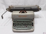Antiga máquina de escrever Remington, na cor cinza. Necessita de revisão.