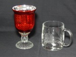 Duas peças em vidro: a) Castiçal em forma de taça, bojo vermelho. Alt. 16cm. b) Caneca gravada com brasão em satiné, Canadá. Alt. 9,5cm.