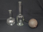 Três peças para laboratório em vidro translúcido, sendo uma garrafa volumétrica, um balão de fundo em cobre redondo (bicado na borda) e um balão de fundo achatado. Alt. 28cm, 21cm e 19cm.