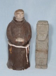 Duas esculturas: a) Esculpida e pedra representando totem de figura tribal. Alt. 18cm b) Em cerâmica policromada moldada na forma de frei. Apresenta leves lascados. Alt. 24cm