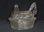 Manteigueira em vidro translúcido moldada na forma de galinha sobre cesto, leve rachado e bicado na borda, tampa adaptada. 12 x 13 x 11cm.
