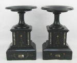 Par de colunas baixas em mármore negro, decoradas com trabalhos de volutas e folhagens em baixo relevo. Detalhes em dourado. Apresentam bicados nas bases e pequenas colagens nos tampos. Alts. 29,5 cm. Diam. tampos 18 cm.