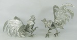 Duas esculturas  em alumínio prateado, representando "Galos" um com uma pata soldada. Meds. 13,5x22x17,5 e 15,5x21,5x17,5cm.