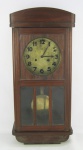 Relógio de parede alemão, com marca da manufatura Junghans. Caixa em madeira entalhada. Porta com vidro. Mostrador em metal, tendo este desgastes. Funcionando, porém necessita regulagem. Med. 64,5x29x15cm.