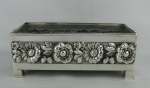 Floreira em metal espessurado a prata, decorada com trabalhos de flores e folhas em alto relevo. Fundo em madeira. Med. 12x31x16 cm.
