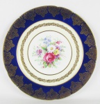 Prato de coleção em porcelana inglesa, com marca da manufatura no verso. Aba no tom azul cobalto e decorado ao centro com pintura floral em policromia. Ricos detalhes em dourado. Diam. 27cm.