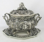 Sopeira com presentoir em faiança européia, com decoração ao gosto Capodimonte, de folhagens, figuras angelicais e volutas em relevo, e cisnes aguisa de alças. Pega da tampa na forma de crianças. Parte interna da sopeira com fio de cabelo. Med. sopeira 31,5x41x24 e Med. presentoir 43x30cm.