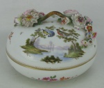 Bela caixa circular em porcelana européia, com marca da manufatura na base. Decorado com pintura em policromia de paisagem com pássaros, lago, flores e folhas. Tampa com alça trabalhada em flores e folhagens em relevo, tendo estas pequenas quebras em algumas folhas. Med. 11x16,5cm.