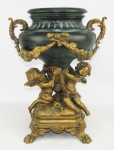 Grande e bela ânfora bojuda em bronze, sendo a base na forma de grupo escultórico dourado, com querubins sobre almofadas com flores, sustentando o vaso. Alças e apliques na forma de volutas e guirlandas. Pés em pata. Med. 40x31x28cm.