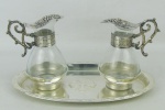 Peça sacra - Par de galhetas com bandeja em metal espessurado a prata, sendo as galhetas em vidro e metal. Bandeja possui ao centro uma cruzem relevo, metal das galhetas com trabalhos de cachos de uva. Med. bandeja 3x22,5x13,5cm.