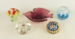 a) Quatro pisa papeis em murano, sendo um ao gosto Millefiori, um na forma de cogumelo (apresenta lascado e batidas), um com flor no interior (apresenta pequena batida interna) e um na forma de fruta.b) Cinzeiro em murano no tom rubi, na forma de peixe, decorado internamente com bolhas de ar. Apresenta lascado na ponta, fissuras e batidinhas internas. Med. 4,5x21x16cm.