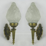 Par de apliques em bronze com trabalhos em gomos, flores e sulcos. Cúpulas na forma de tocha em vidro fosco, uma apresentando quebra na parte interna do encaixe, somente visível quando retirada. Alt. total 46cm.