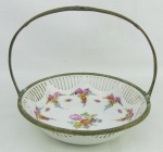 Petisqueira na forma de cesta, em porcelana alemã, marca da manufatura Schumann, decorada com pintura flora. Borda e alça em metal ormulu, com flores e folhas. Med. 21x22 cm.