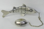 Coador para chá, na forma de ovo, tendo este como suporte de figura de peixe em prata com contraste de teor 833 milésimos. O coador é em metal espessurado a prata. Peso do peixe 65 g. Comp. total com a corrente 35 cm.