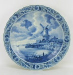 Medalhão em porcelana Holandesa, com marca da manufatura Delft no verso, decorado com paisagem com moinho de vento em tons de azul. Parte de trás da borda com dois lascados perceptíveis apesar de ser no verso. Diam. 39 cm.