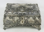 Bela caixa em metal espessurado a prata, possivelmente europeia, com marca da manufatura na base. Profusamente decorada com trabalhos em relevo de flores, folhagens, pássaros e paisagem com casas. Parte interna forrada com tecido vermelho. Desgastes no prateado. Med. 12,5x24x15cm.