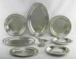 Baixela em metal espessurado a prata, marca da manufatura Magna Prata, composta de 3 travessas ovaladas, 2 redondas, molheira e terrina com tampa, esta reversível para travessa funda. Marcas de uso. Med. maior 39,5x24 cm.