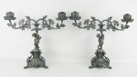 Par de candelabros para 2 velas, em metal patinado, sendo sustentados por figuras de Puttinos sobre bases triangulares com pés em volutas. Braços com trabalhos de cachos de uva e parreiras. Med. 36,5x44x15cm.