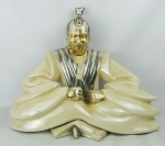 Estatueta em faiança no tom creme, com detalhes prateados e dourado, representando "Figura oriental em meditação". Dourado e prateado com desgastes, com pequena perda no esmalte atrás de um braço. Med. 39x51x27cm.
