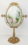 Espelho de mesa em bronze dourado, trabalhado com flores e folhagens em relevo. Espelho oval com regulagem de inclinação. Apresenta mancha do tempo. Verso com placa em porcelana decorada com pintura floral em policromia. Alt. total 44,5cm.