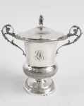 REINO DA SARDENHA, CIDADE DE GENOVA RICA TAÇA COBERTA EM PRATA DE LEI BATIDA, REPUXADA E CINZELADA. MARCAS PARA O ANTIGO REINO DA SARDENHA E CIDADE DE GÊNOVA.  PEGA COM FEITIO DO DEUS LUPÉRCIO TOCANDO SUA GAITA. ALÇAS LATERAIS QUE SE ELEVAM DECORADAS COM LEOPARDOS. TERÇO INFERIOR COM LINDO MOSAICO GEOMÉTRICO RELEVADO. ITÁLIA, PRIMEIRA METADE DO SEC. XIX. 20 CM DE ALTURA. 375 GNOTA: Pã  para os gregos e Lupércio para os romanos, na mitologia grega, é o deus dos bosques, dos campos, dos rebanhos e dos pastores. Vive em grutas e vaga pelos vales e pelas montanhas, caçando ou dançando com as ninfas. É representado com orelhas, chifres e pernas de bode, amante da música, traz sempre consigo uma flauta. É temido por todos aqueles que necessitam atravessar as florestas à noite, pois as trevas e a solidão da travessia os predispõem a pavores súbitos, desprovidos de qualquer causa aparente, e que são atribuídos a Pã; daí o termo "pânico". Pã apaixonou-se pela náiade Sírinx, que rejeitou com desdém o seu amor, recusando-se a aceitá-lo como seu amante pelo fato de ele não ser nem homem, nem bode. Pã então perseguiu-a, mas Sírinx, ao chegar à margem do rio Ladon e vendo que já não tinha possibilidade de fuga, pediu às ninfas dos rios, as náiades, que mudassem a sua forma. Estas, ouvindo as suas preces, atenderam ao seu pedido e a transformaram em caniço. Quando Pã a alcançou e quis agarrá-la, não havia nada, exceto o caniço e o som que o ar produzia ao atravessá-lo. Ao ouvir aquele som, Pã ficou encantado e resolveu então juntar caniços de diferentes tamanhos, inventando um instrumento musical ao qual chamou Siringe, em honra à ninfa. Esse instrumento musical é mais conhecido pelo nome de flauta de pã, em honra ao próprio deus. Pã teria sido um dos filhos de Zeus com sua ama de leite, a cabra Amalteia. Seu grande amor no entanto foi Selene, a Lua.