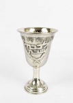 HAZORFIN  BELO COPO PARA KIDUSH EM PRATA DE LEI. DECORADO COM CACHOS DE UVA, INSCRIÇÕES EM HEBRAICO E LAUREL. ISRAEL, SEC. XX. 12 CM DE ALTURANOTA:  A Torá refere-se a dois requisitos relativos ao Shabat - "mantê-lo" e para "lembrar-lo" (shamoreZakhor).A lei judaica exige, portanto, que o Shabat seja observado em dois aspectos. É preciso "mantê-lo", abstendo-se de trinta e nove atividades proibidas, e deve-se "lembre-se que" ou rememorá-lo fazendo-se arranjos especiais para o dia, e, especificamente, através da cerimônia de kiddush. Recitar kiddushantes da refeição na véspera do Shabat e feriados judaicos é, portanto, um mandamento da Torah (como é explicado pela Torá Oral).Para honrar amitzvah deve-se recitar a prece kiddush sobre o copo de vinho. Para tanto, uma taça de prata é usada frequentemente. O ritual pressupõe realizar uma revi'it de líquido (um quarto do copo medido com polegar).A revi'ité entre 161,5 ml e 90,7 ml (de acordo com o tamanho do copo). Depois que a pessoa recita a bênção e bebe do copo, o vinho é passado ao redor da mesa ou derramado em copos pequenos para os outros participantes.