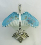 Belo centro de mesa em metal, possivelmente alemão da manufatura W. M. F., decorado com folhagens e volutas em relevo. Prato em opalina no tom azul e leitoso, decorado com pintura floral em policromia, e trabalhado com babados. Med. 28x23,5x28,5cm.