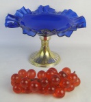 Centro de mesa em metal espessurado a prata, com prato em vidro no tom azul cobalto, com borda trabalhada em babados. Acompanha peça decorativa em acrílico representam cachos de uvas. Metal com desgastes no prateado. Med. centro de mesa 18x28,5cm.