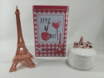Lote contendo 3 itens decorativos representando Paris: 1 torre Eiffel em tom rosé, 1 lata porta objetos retangular e 1 pequeno potiche decorado pela palavra `Paris` em sua pega. Maior tamanho 19 cm.