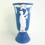 JASPER WARE - Vaso em biscuit Europeu, na tonalidade azul com ornamentação no estilo neoclássico na cor branca, obtida através da técnica Pate-sur-pate, representando figura feminina grega segurando cacho de uva e cálice, na parte posterior do corpo coroa de flores suspensa por laçarote encimado por folhagem, base abaulada cercada por elementos fitomorfos em relevo. Exemplar em excelente estado. Dimensões: 15 x 8 cm.