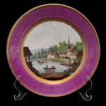 Prato em porcelana possivelmente francesa. Pasta dura. Borda com larga barra na cor bordeaux circundada por friso em ouro. Centro com paisagem, apresentando montanha, rio, casas e personagens. França. Séc. XIX. 20,5 cm.