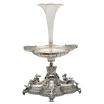 Belíssimo centro de mesa de formato oblongo em prata alemã repuxada e cinzelada, profusamente ornamentado por adornos e estilizações, centralizando floreira com tulipa em cristal. Base lobulada ornada por frisos estilizados, realçada por figuras femininas em relevo nas extremidades e medalhões com putti nas laterais. Parte superior com pedestal lavrado sustentando recipiente ovalado moldado com gomos. Pés recurvos no formato de folhas. Contraste da prata alemã de teor 800 mls e marca do prateiro Gebruder Kuhn. Circa de1890/1900.  63 x 43 cm. Peça adquirida do acervo do Antiquário Nobrega. Centro de mesa de prata alemã com floreiro de cristal. 63 cm / 43 cm.