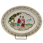 Travessa oval em porcelana Companhia das Índias. Apresenta decoração em esmaltes policromados com larga borda florida e centro com figura em jardim. China. Dinastia Qing (1644-1911). Reinado Qianlong (1736-1795).  29 x 37 cm.